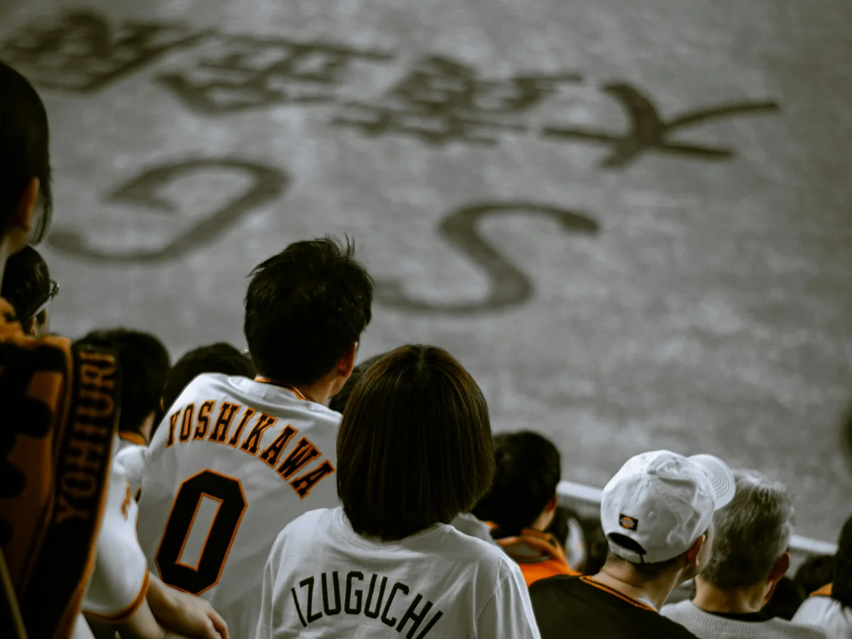 Tifosi con le maglie bianche e arancioni dei Yomiuri Giants, visti di spalle, che seguono la partita dai loro posti