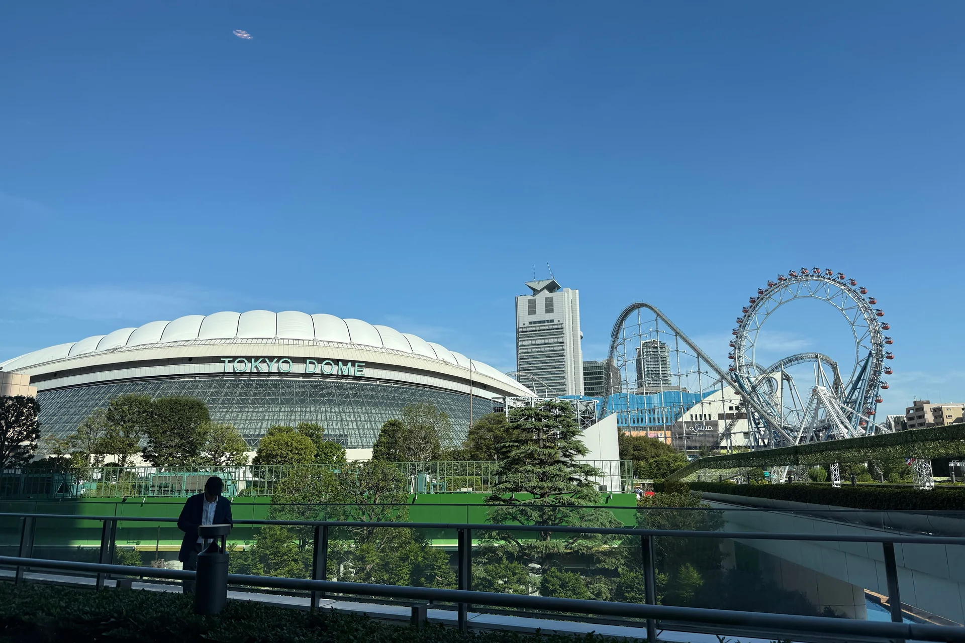 Det hvide luftbårne tag på Tokyo Dome med påskriften 'TOKYO DOME', fotograferet i dagslys udefra stadionet, med forlystelsesparken Tokyo Dome City synlig bagved